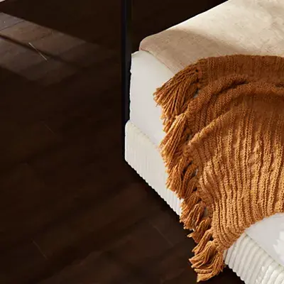 Brown fringed blanket on a bed over dark hardwood flooring.