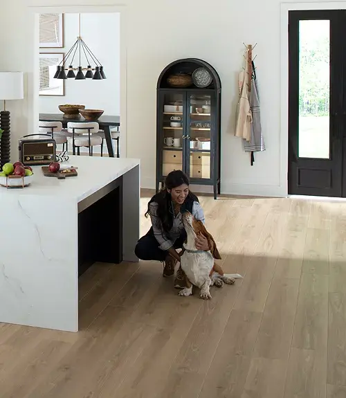 Woman petting a basset hound on light wood-look flooring in a kitchen.