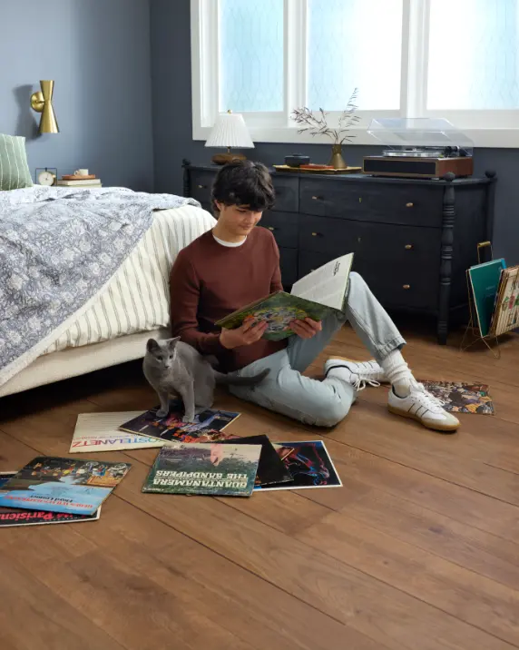 Medium brown wood-look flooring in a bedroom with a young man and grey cat sitting on the floor with vinyl records.