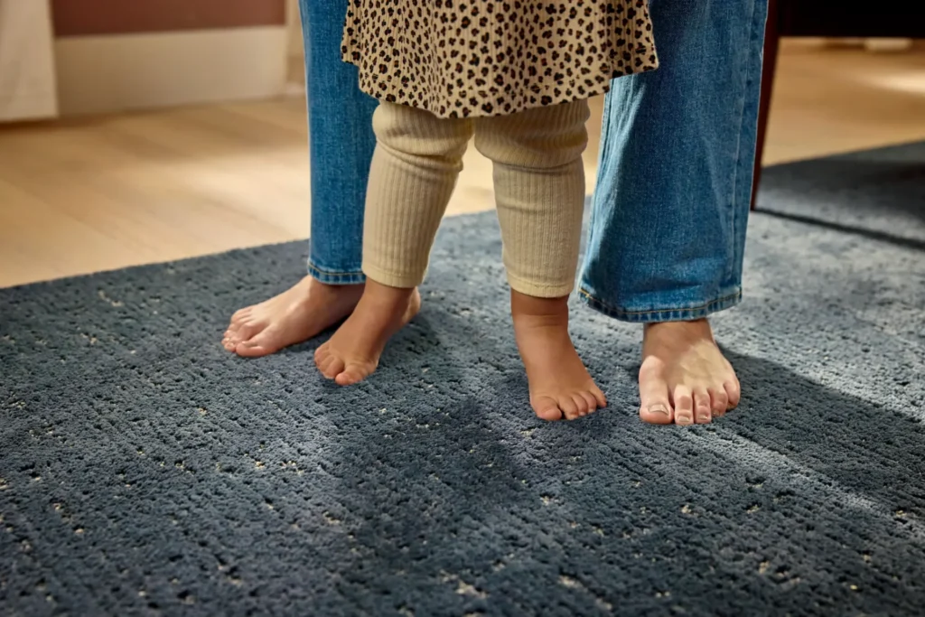 Bare feet of a toddler and adult standing on a plush, textured blue carpet.