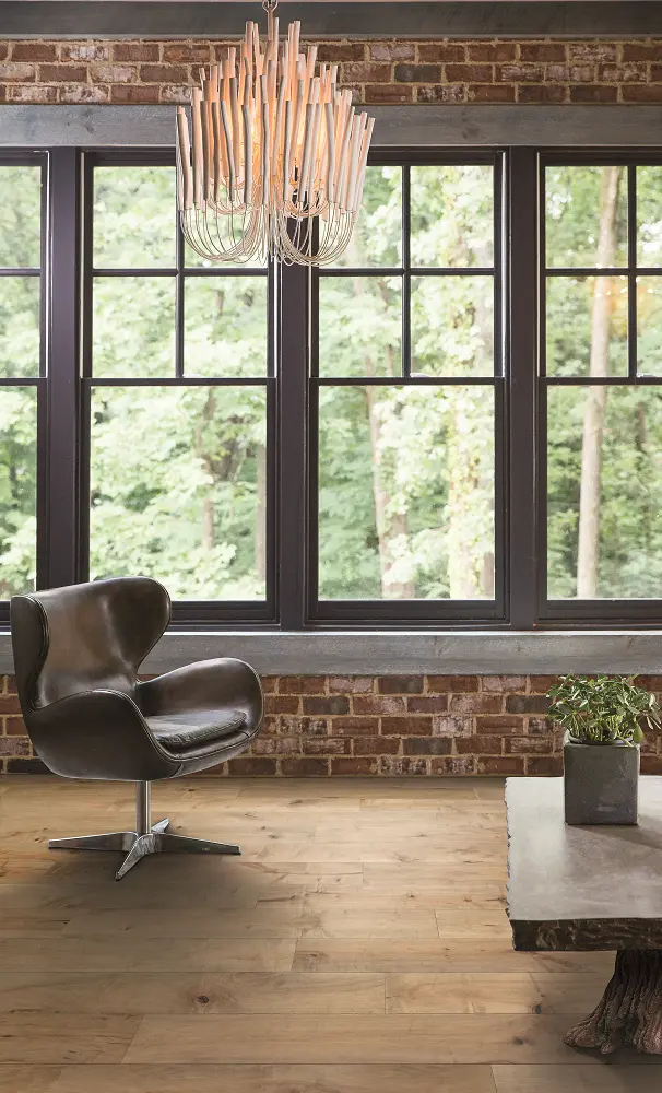 Light oak hardwood flooring in a room with large dark windows and a exposed brick wall.