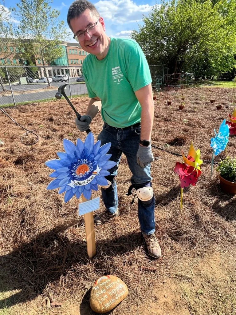 YMCA Garden - Bruce: Man smiling with a hammer while installing a decorative flower sign in a garden.