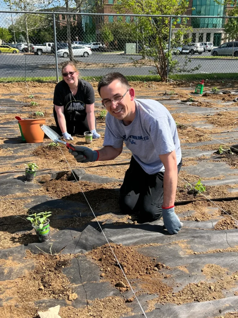 YMCA Garden - Kimberly & Joe: Man and woman smiling while planting seedlings in an outdoor garden plot.